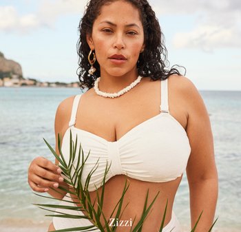 Kvinde med krøllet mørkt hår iført hvid bikini-top og skallenæppe, der holder et grønt palmeblad ved havet med et fjernt kystlandskab.