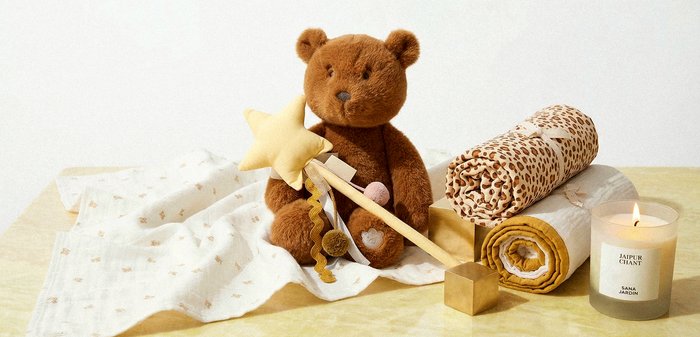 Καφέ αρκουδάκι που κρατάει μια απαλής υφής ράβδο με αστέρι, περιτριγυρισμένο από υφασμάτινες κουβέρτες με σχέδια, έναν χρυσό κύβο και ένα αναμμένο αρωματικό κερί σε μια ανοιχτόχρωμη επιφάνεια.