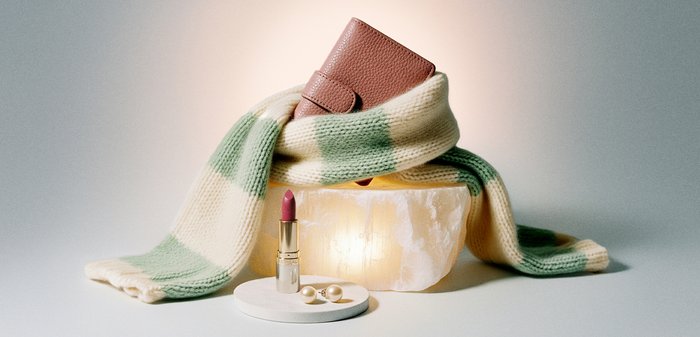 Braune Lederbrieftasche, eingewickelt in einen grün-weißen Strickschal, auf beleuchtetem Stein, mit pinkem Lippenstift und Perlenohrringen auf weißem Tablett.