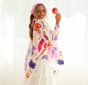 Bunt bedruckter Kimono mit langen Ärmeln, aus leichtem Stoff. Über einem weißen Kleid getragen, mit einem rosa Blumenaccessoire befestigt.