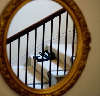 Chaussures noires avec des sangles blanches posées sur des escaliers en moquette beige, vues à travers un miroir rond avec un cadre doré, avec des rampes en bois en vue.