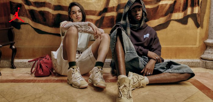 Deux jeunes sont assis par terre contre un mur à motifs, tous deux portent des baskets beige et des vêtements de ville décontractés ; le logo rouge Jordan est visible en haut à gauche.