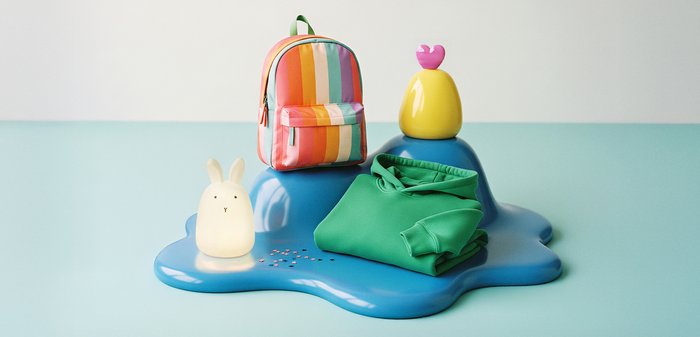 Opgevouwen groene hoodie, rugzak met regenboogstrepen, verlichte konijnenlamp en geel voorwerp met roze hart op blauwe abstracte ondergrond.