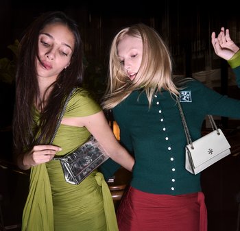 Två personer visar upp väskor: en silverfärgad metallisk kuvertväska med kedjerem och en liten vit handväska med logotyp, mot en mörk bakgrund.