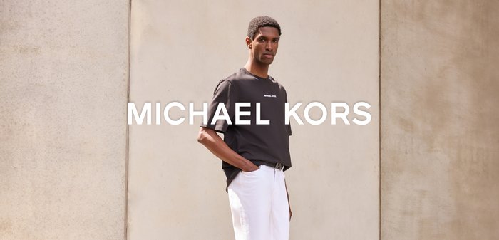 Mężczyzna ubrany w czarną koszulkę Michael Kors i białe spodnie stoi przy beżowej ścianie z logo Michael Kors na środku.