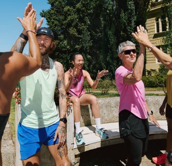 Gruppe von Menschen in Sportbekleidung, mit verschiedenen Farben: mintgrünes Tanktop, pinkes Outfit und schwarze Shorts, die draußen High-Fives geben.