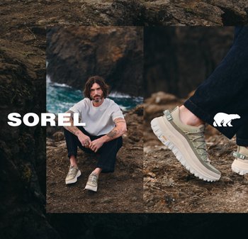 Man with tattoos sitting on rocky coastline wearing beige hiking shoes and black pants, zoomed-in shot of shoe sole and detailing.