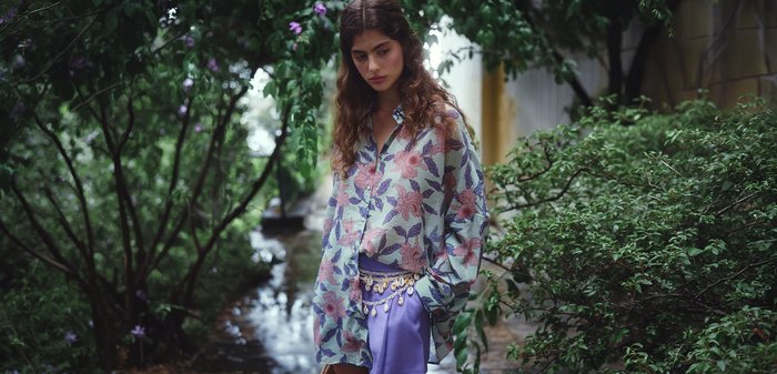 Giovane donna con lunghi capelli ricci indossa una camicia floreale e pantaloncini viola con una cintura di conchiglie, in piedi vicino a piante verdi rigogliose e fiori.