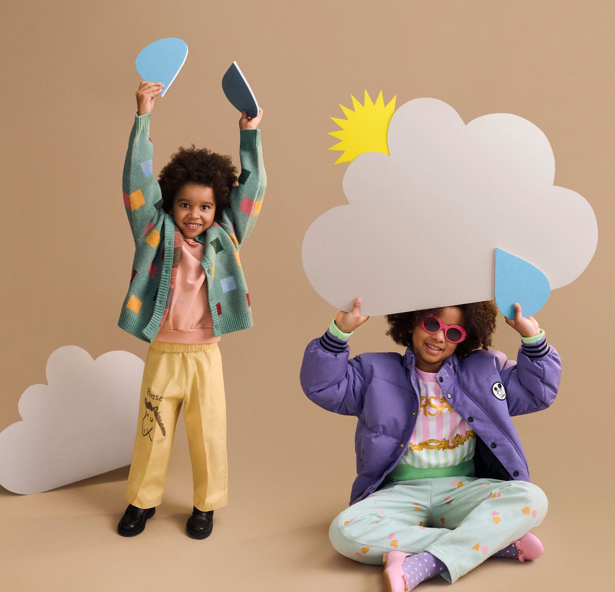 Kaks last mängivad ilmateemaliste rekvisiitidega; üks seisab hoides vihmapiisa kujutisi, teine istub ja kannab päikeseprille pilve lõike all.