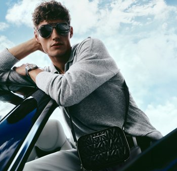 Ung mann med solbriller og grå genser, lent mot en bildør, med en svart Michael Kors skulderveske og en sølvklokke.