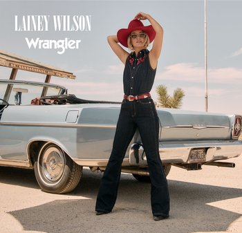 Kvinne i mørk denimdresser og rød hatt poserer ved en vintage kabrioletbil på en solrik dag med klar himmel.