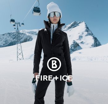 Czarna kurtka narciarska z zamkiem, wysokim kołnierzem i dopasowanym fasonem. Białe rękawice trzymane w rękach, zaśnieżone górskie tło, w oddali wyciąg narciarski. Wyeksponowane logo marki.