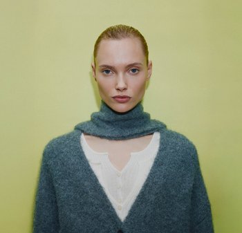 Graue, weiche Wolljacke mit hohem Kragen, kombiniert mit einem weißen Button-up-Oberteil. Der Hintergrund ist ein einheitliches, hellgelbes.