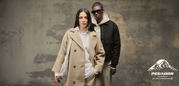 Palton bej cu două rânduri de nasturi, hanorac gri și pantaloni bej purtați de o femeie; jachetă bomber neagră peste hanoracul gri și pantaloni bej purtați de un bărbat. Ochelari de soare negri și pantofi.