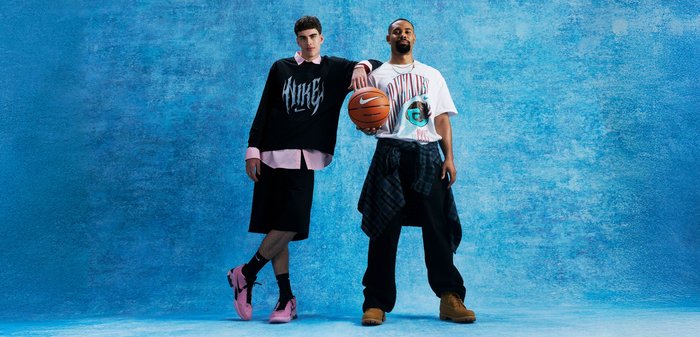 Two men in casual streetwear pose against a blue backdrop, one holding a basketball and the other leaning on his shoulder.