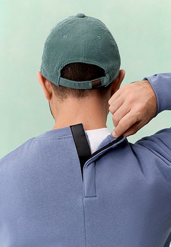 Person mit grüner Kappe stellt den Reissverschluss am Rücken eines blauen Sweatshirts vor einem einfarbigen hellgrünen Hintergrund ein.