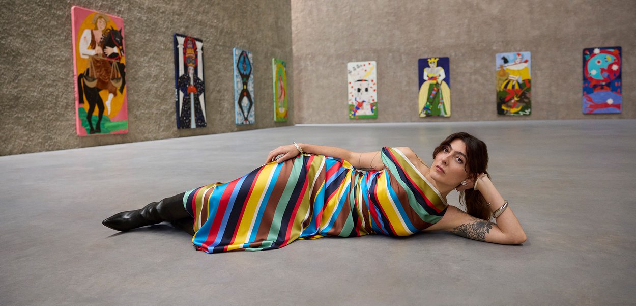 Värvikas triibuline kleit satiinkangast; horisontaalsed triibud punases, sinises, rohelises, kollases ja pruunis; mustad põlveni ulatuvad saapad; minimalistlik galerii keskkond.