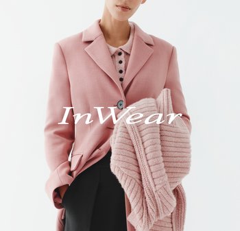 Person trägt ein pinkes Sakko und ein gestricktes pinkes Oberteil, hält einen gefalteten, gerippten pinken Pullover vor einem schlichten weißen Hintergrund.