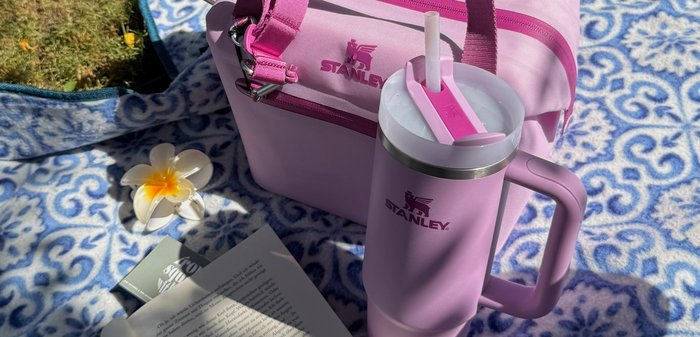 Rosafarbener Stanley Isolierbecher mit Strohhalm und passender Lunchbag auf blau gemustertem Decke, neben offenem Buch, Brille und weißer Blume.