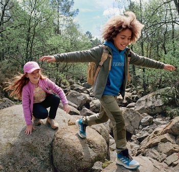 Due bambini con abiti casual e zaini che fanno escursioni su un terreno roccioso in una foresta, uno che si equilibra su una roccia e l'altro che si accovaccia.