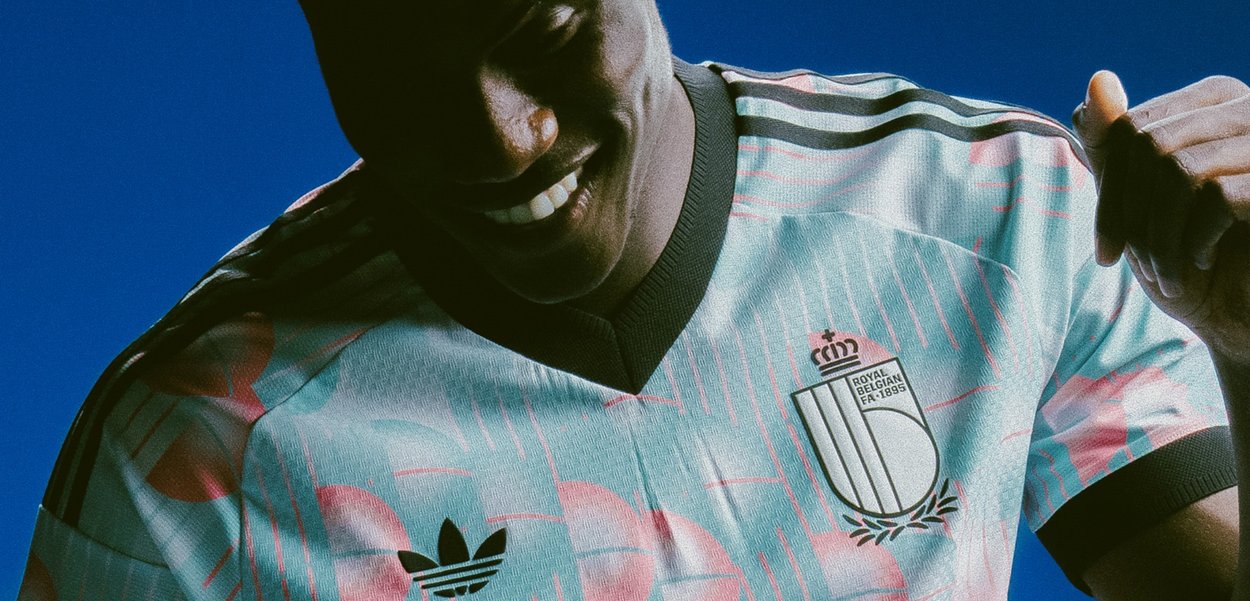 Personne portant un maillot de football Royal Belgium bleu et rose avec des logos Adidas, souriant et levant le poing sur un fond bleu.