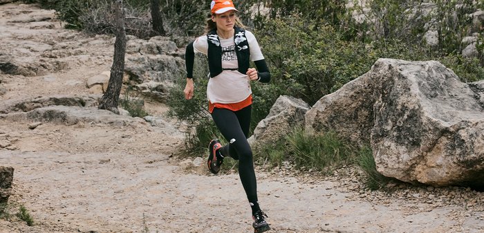 Loper met oranje pet en zwarte legging joggend op een rotsachtig pad omringd door bomen en grote rotsblokken.