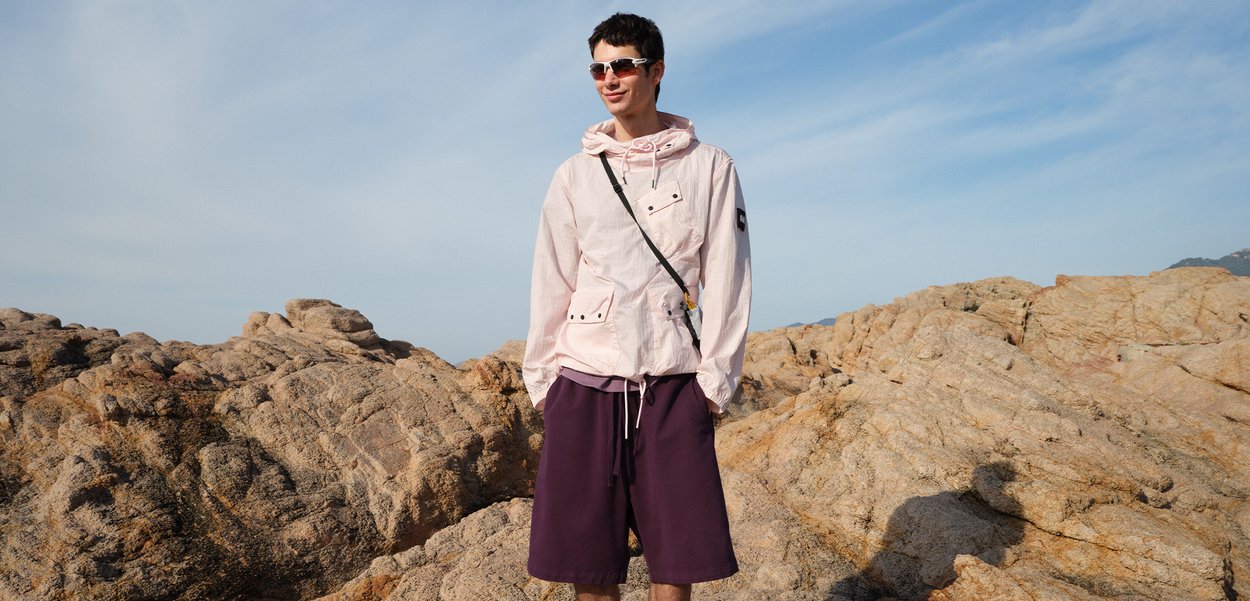 Hombre con gafas de sol, chaqueta rosa claro y pantalones cortos morados de pie sobre un terreno rocoso bajo un cielo azul nublado.