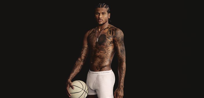 Athletischer Mann mit geflochtenen Haaren und Ganzkörpertätowierungen hält einen Basketball, trägt weiße Calvin Klein Boxershorts vor schwarzem Hintergrund.