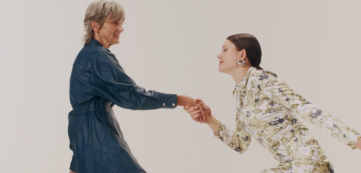 Două modele se țin de mână, una purtând o rochie cămașă din piele albastră închisă cu talia strânsă, cealaltă într-o rochie ajustată cu model floral.