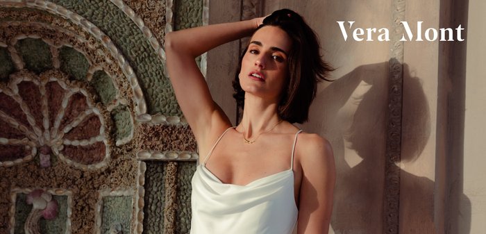 Frau in einem weißen Spaghettiträgerkleid posiert mit einem erhobenen Arm vor einer kunstvollen Steinmauer, während Sonnenlicht Schatten wirft.