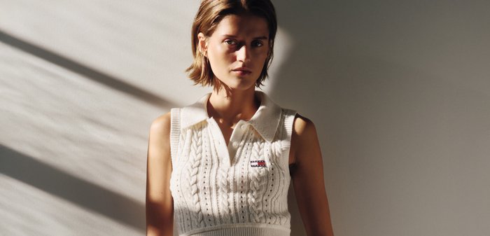 Mujer con cabello corto y castaño que lleva un polo sin mangas de punto blanco con un pequeño logo, de pie frente a una pared beige con patrones de luz y sombra.