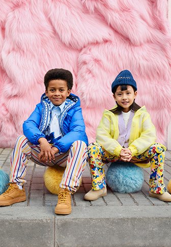 Duas crianças sentam-se em pufes macios e coloridos. Uma veste uma jaqueta acolchoada azul, calças às riscas e sapatos castanhos; a outra usa uma jaqueta amarela, calças florais e um gorro azul. Um fundo rosa e peludo é visível.