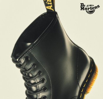 Zwarte leren Dr. Martens laars met gele stiksels, zwarte veters en merklabel aan de treklus, afgebeeld tegen een lichte achtergrond.