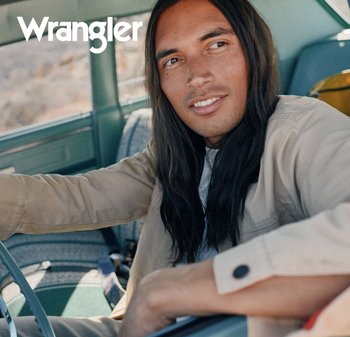 Mann med langt mørkt hår iført en beige jakke som sitter i en veteranbil og holder rattet, Wrangler-logo øverst til venstre.