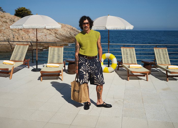 Homme avec des lunettes de soleil et des tatouages se tient sur la terrasse au bord de la piscine près des chaises longues et des parasols au bord de la mer, tenant un sac tissé.