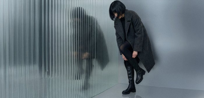 Femme portant un manteau sombre et des bottes ajuste sa botte en se tenant à côté d'un mur en verre givré avec son reflet visible.
