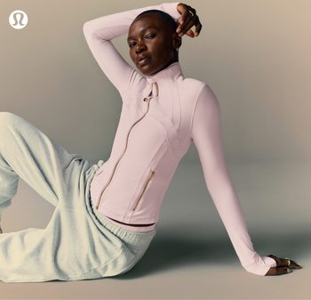 Donna in posa seduta sul pavimento, indossando una giacca leggera rosa chiaro con zip e pantaloni della tuta grigio chiaro, su uno sfondo neutro.