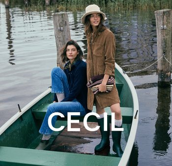 Twee vrouwen op een groene boot bij het water. De ene draagt een donker corduroy jurk; de andere draagt een blauwe blouse en denim jeans, met rubberen laarzen.