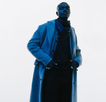 Langer grauer Wollmantel über einem schwarzen Hemd, mit einem Muster aus Knöpfen und einem dunklen Schal. Glatte Textur, mit einem lässigen Schnitt und dunklen Hosen.