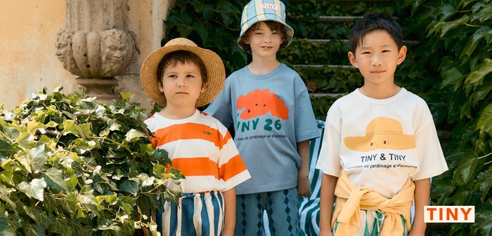 Tre bambini con abiti estivi e cappelli sono in piedi su gradini coperti da tessuto a righe circondati da fogliame verde.