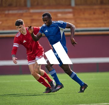 Deux joueurs de football masculins sur un terrain vert ; l'un portant un maillot rouge avec des rayures blanches, l'autre un maillot bleu avec des accents blancs.