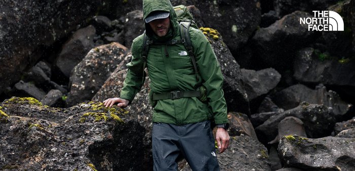 Grüne wasserdichte Jacke mit Kapuze, verstellbarem Gürtel und Logodetail. Kombiniert mit grauen wasserdichten Hosen, vor einer felsigen Landschaft mit Feuchtigkeit.