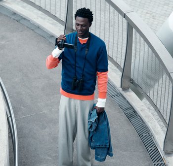 Mann in einem mehrlagigen blauen und orangefarbenen Oberteil hält eine Videokamera in der einen Hand und eine blaue Jacke in der anderen, steht auf einem geschwungenen Fußweg.