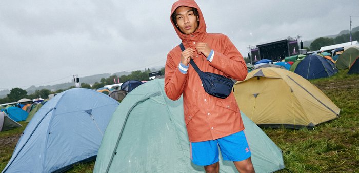 Personne en imperméable orange et short bleu ajustant sa capuche, debout parmi des tentes sur de l'herbe mouillée lors d'un événement en plein air par temps nuageux.