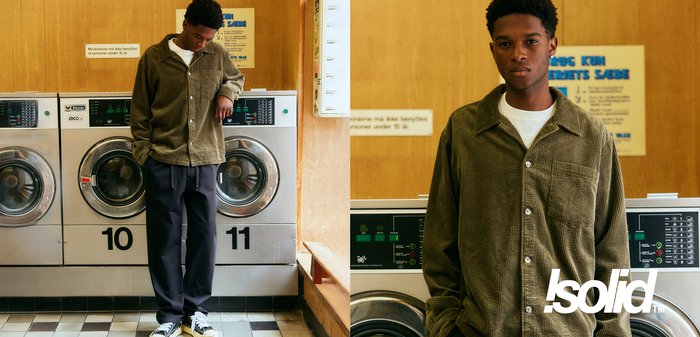 Olivgrünes Cordhemd mit Knopfleiste und Brusttasche; kombiniert mit dunklen Hosen und schwarzen Sneakers, steht neben silbernen Waschmaschinen.