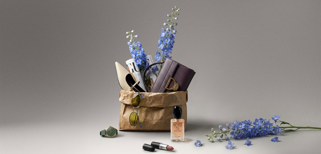 Braune Papiertüte mit einer Handtasche, weißen High Heels, Sonnenbrille, eingerolltem Magazin und blauen Blumen, daneben Uhr, Lippenstift, Parfüm und Blumen.