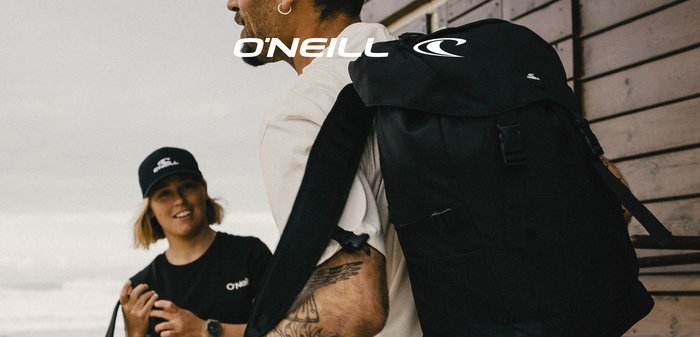 Man met een getatoeëerde arm en een grote zwarte rugzak, pratend met een vrouw in een zwart O'Neill-shirt en pet, staand bij een houten muur op een strand.