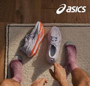 Lichtpaarse sport schoenen met oranje accenten en gestructureerde zolen naast een voet in een roze met patroon sok op een gestructureerd tapijt.
