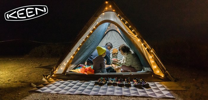 Family of four inside a lit triangular tent at night, sitting on a mat with colorful shoes lined up outside.