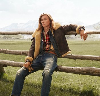 Giacca foderata in shearling marrone sopra una camicia di denim blu, flanella a quadri rossi e jeans blu strappati. Dettagli di guanti e orologio visibili.
